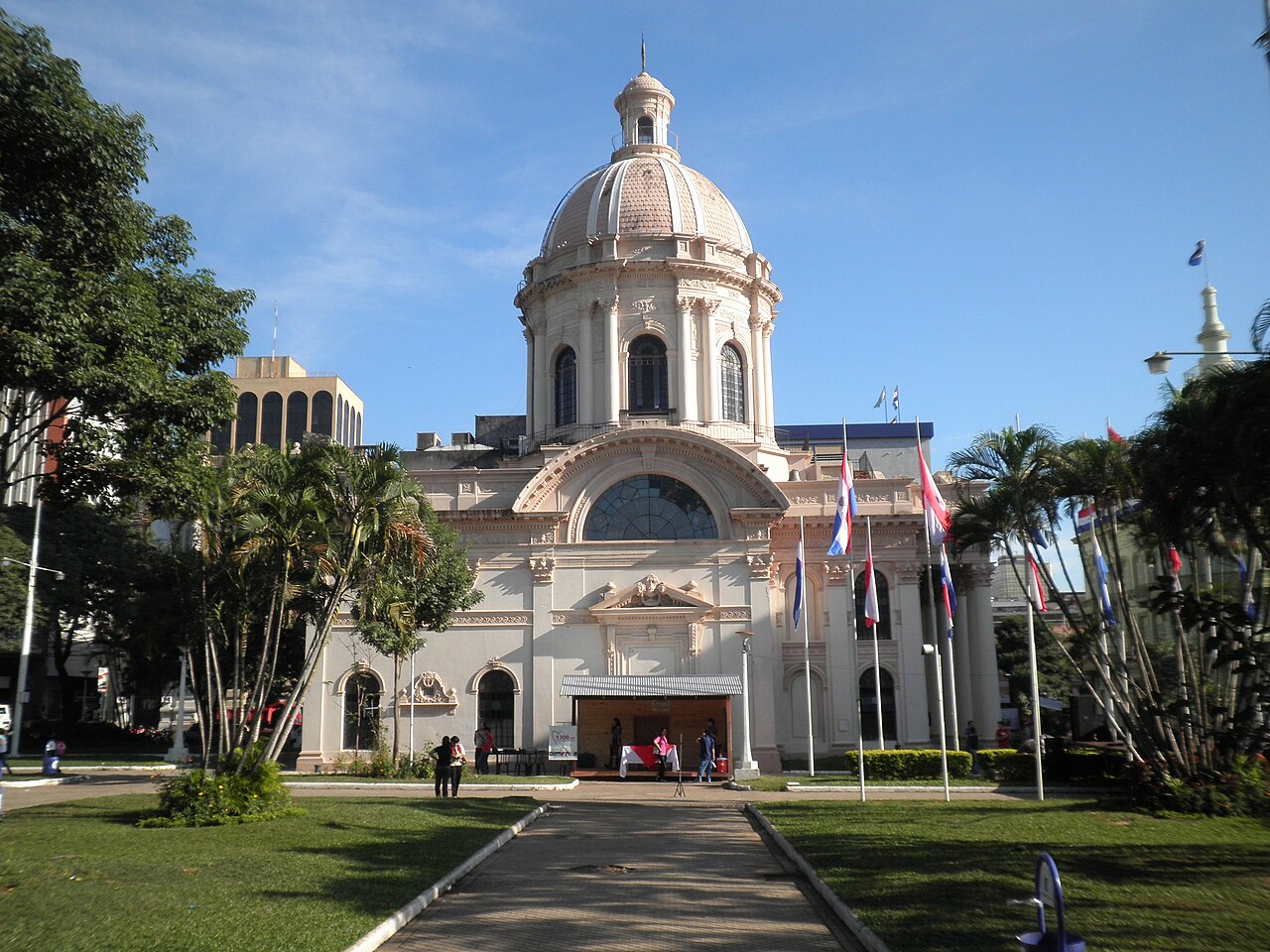 Domed façade of the National Pantheon of the Heroes in Asunción, with flags, palms and a path across the foreground.