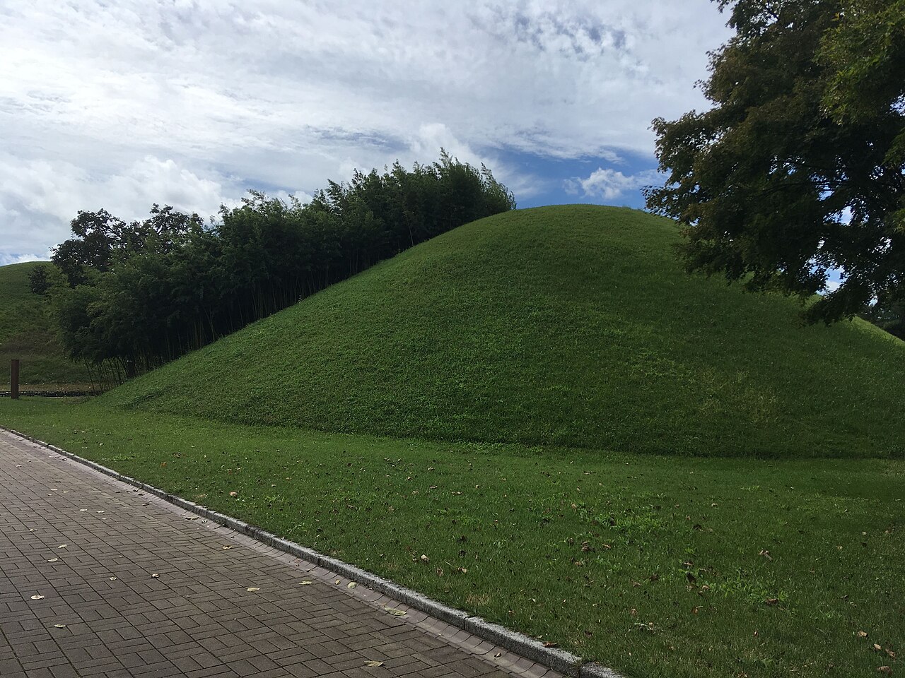 A grass-covered burial mound beside a paved path and trees at Daereungwon in Gyeongju.