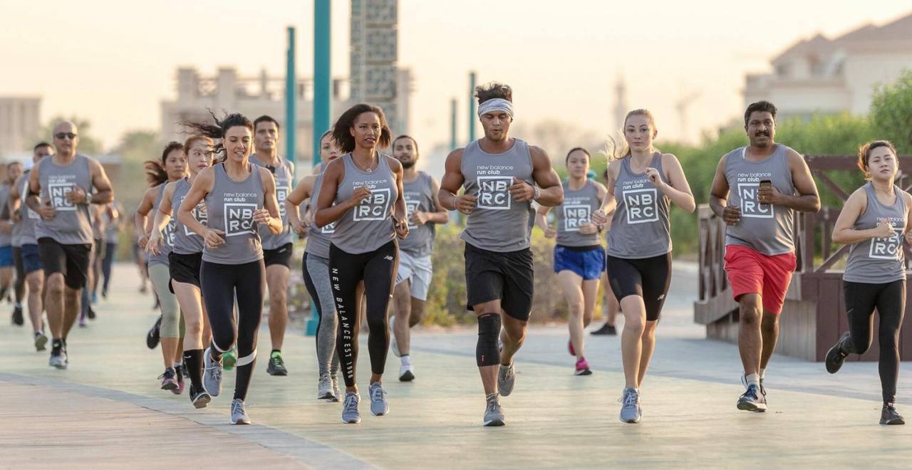 A group of runners wearing grey New Balance Run Club tops jogging along an urban promenade.