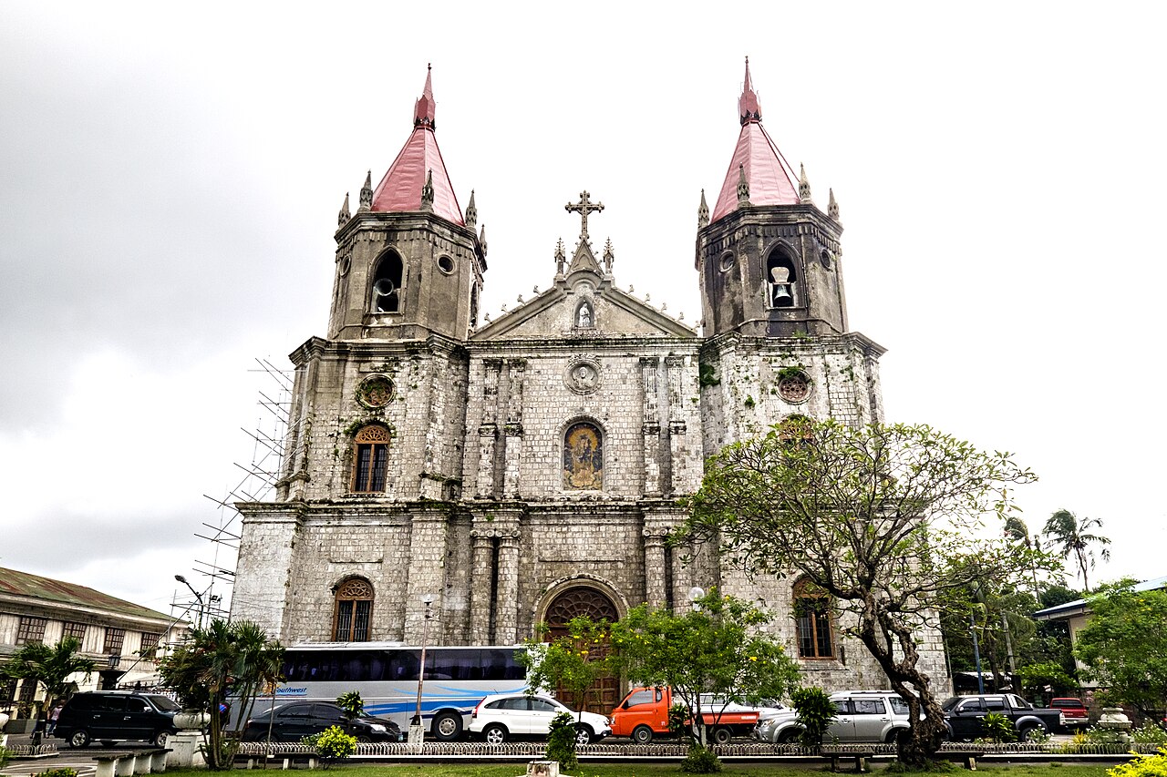 Stone façade and twin red spires of Molo Church in Iloilo City, with trees and vehicles in front.