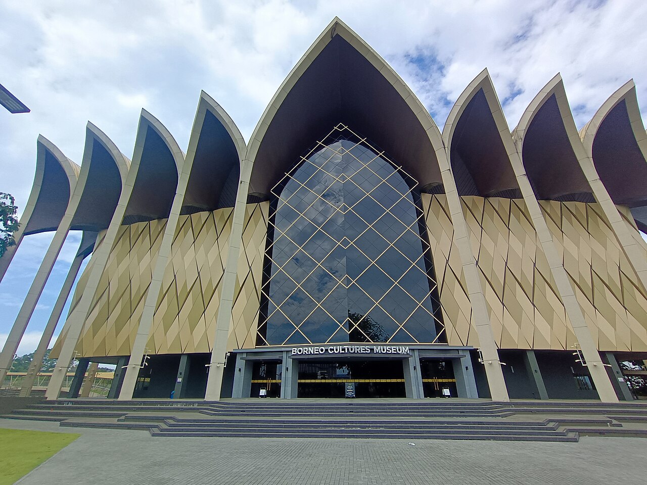 Gold-toned peaked façade and central glass entrance of the Borneo Cultures Museum in Kuching, viewed from the forecourt.