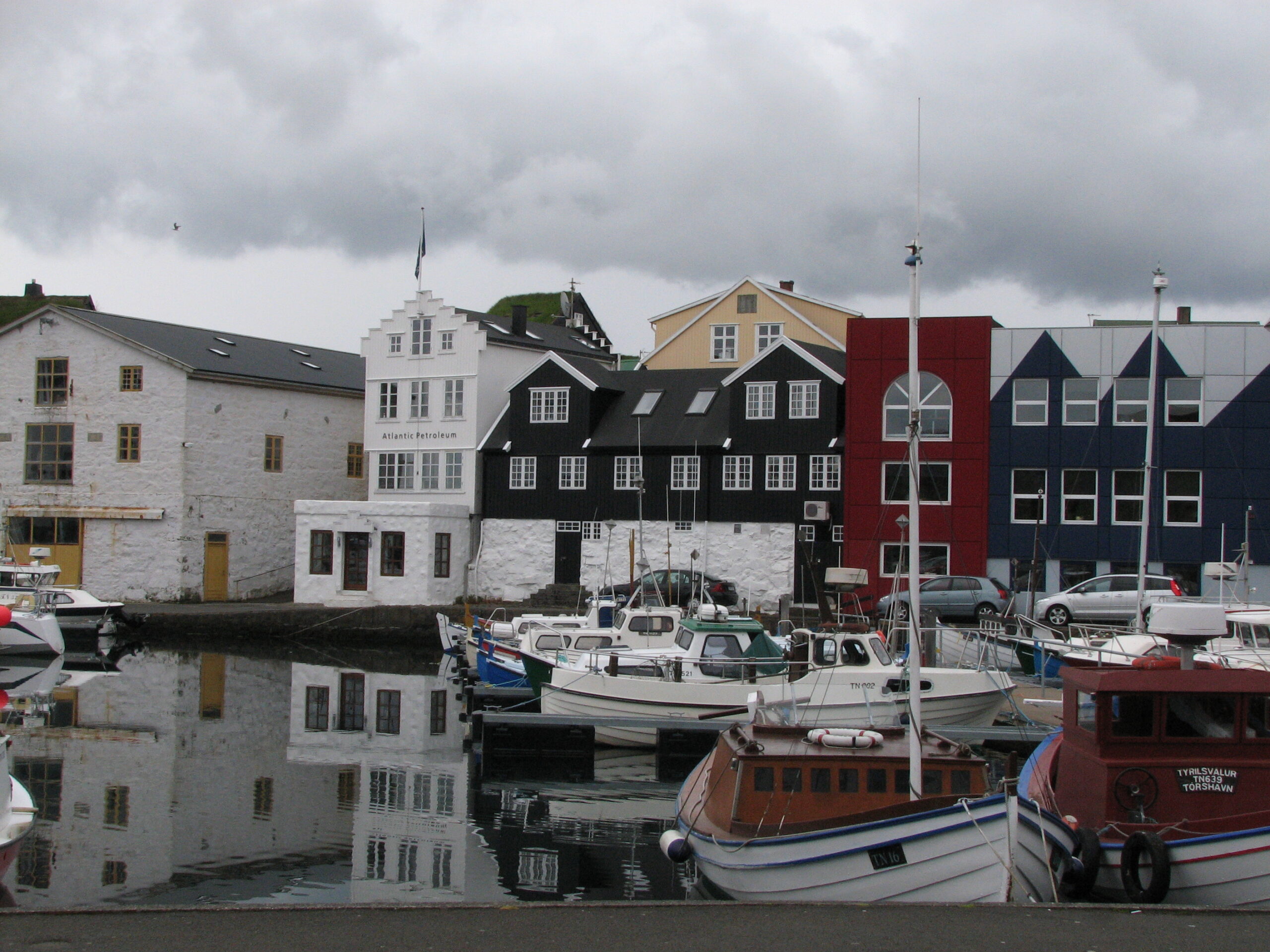 Boats reflected in Tórshavn harbour beneath colourful waterfront buildings and a cloudy sky.