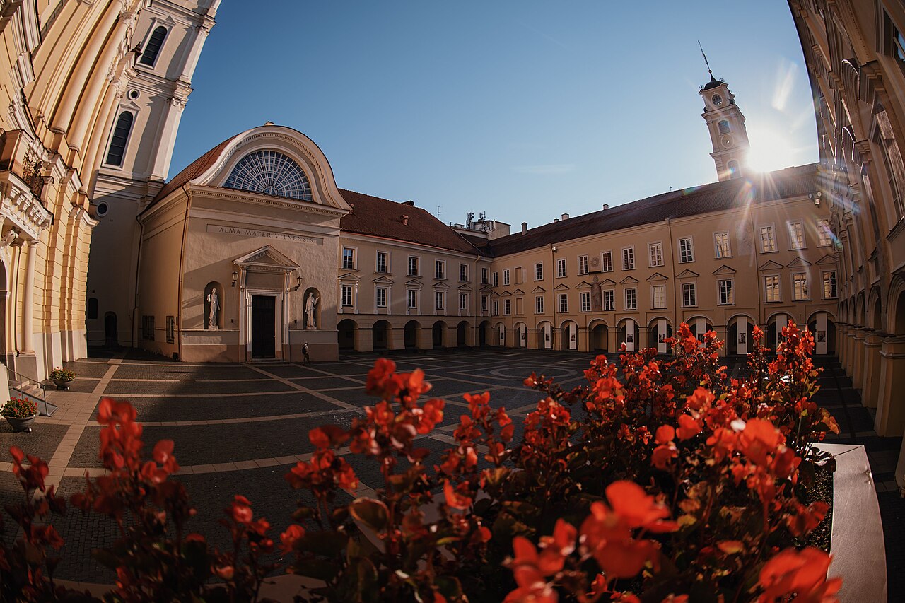 Vilnius University’s Grand Courtyard with arcaded buildings, a church façade and red flowers in the foreground.